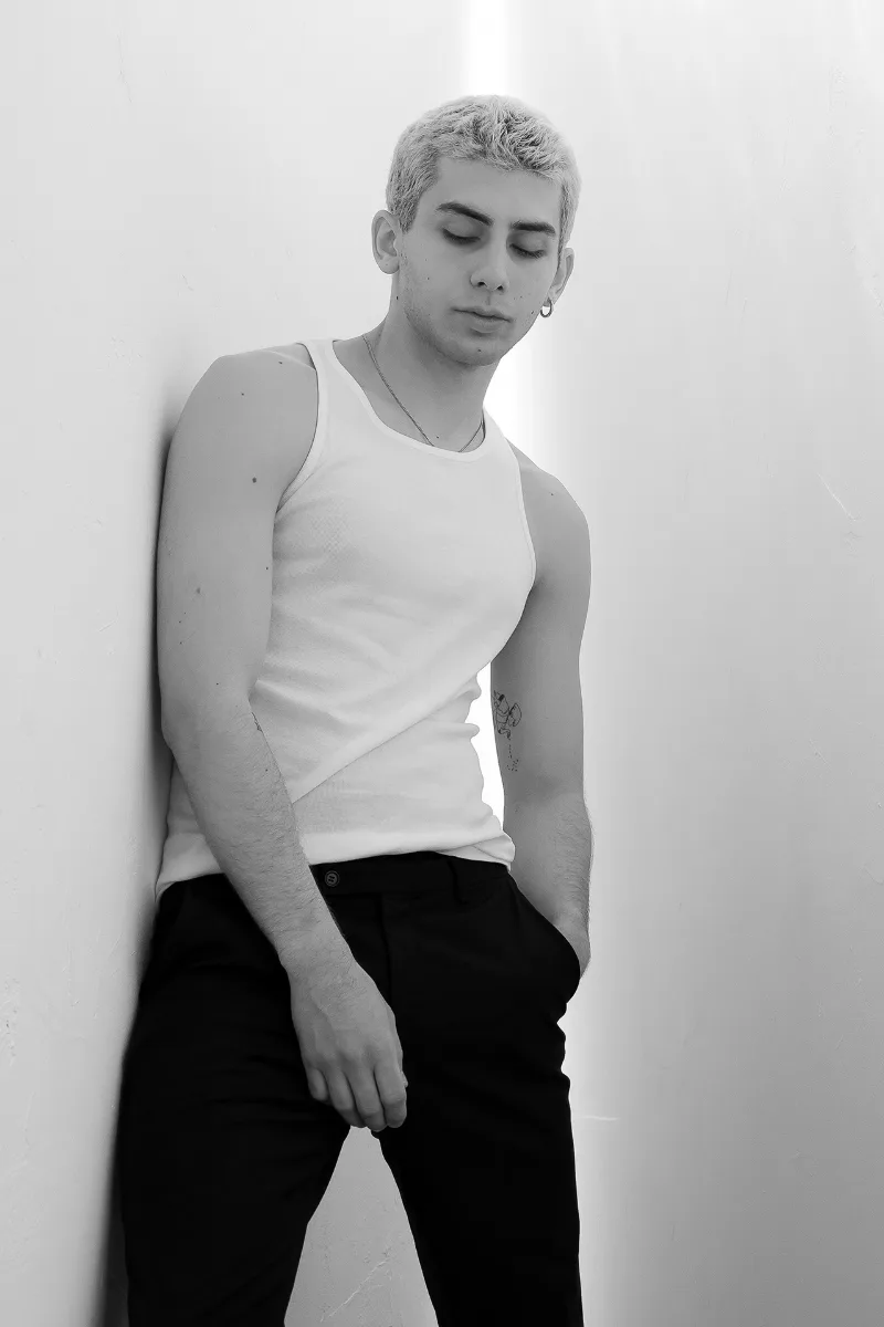 Schwarz-Weiß-Beauty-Portrait eines jungen Mannes mit hellem Haar, weißes Tanktop, an eine Wand gelehnt