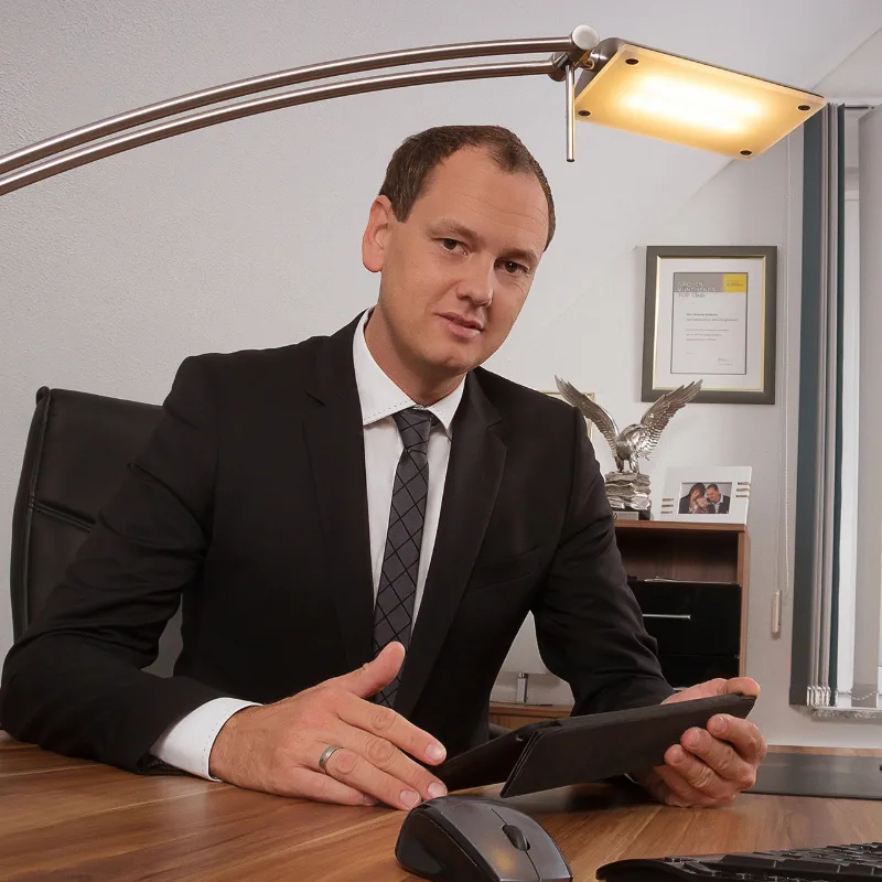 Business-Portrait: Mann im dunklen Anzug mit grauer Krawatte am Schreibtisch, in der Hand ein Tablet, Schreibtischlampe im Vordergrund