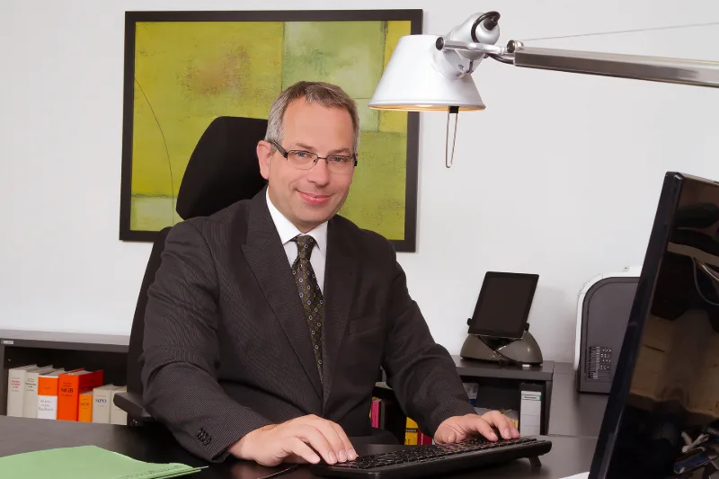 Geschäftsmann im Anzug am Schreibtisch, im Hintergrund Aktenrücken und ein modernes Wandbild