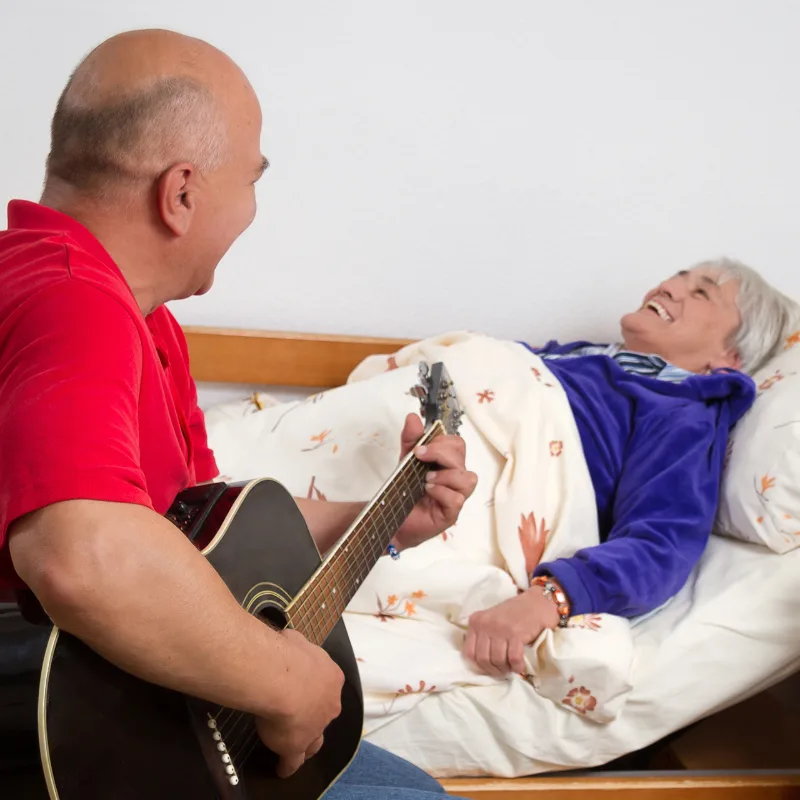 Pfleger mit roter Polo-Shirt spielt einer lachenden Patientin im Bett auf der Akustikgitarre vor
