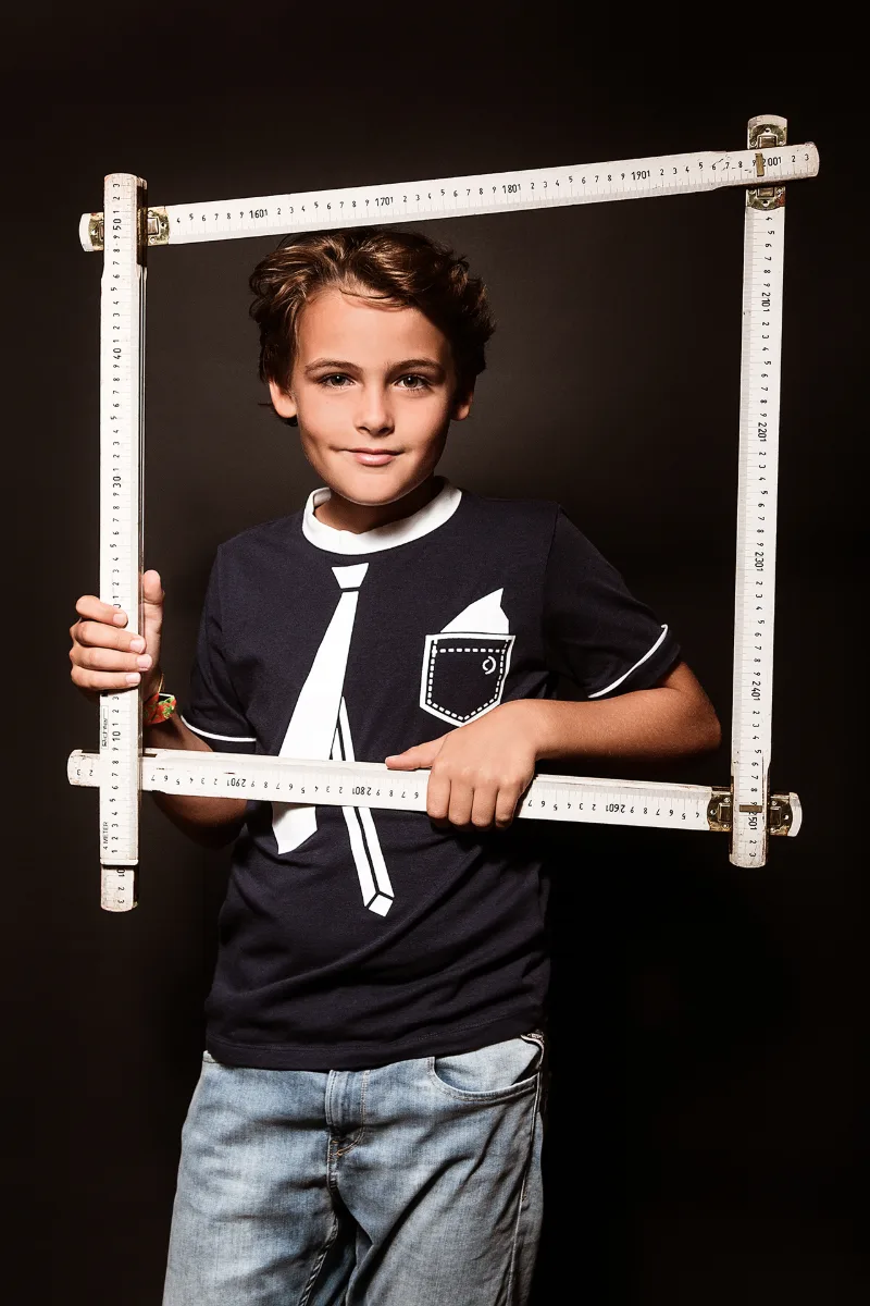Junge rahmt sein Gesicht mit einem zum Quadrat gefalteten Zollstock vor schwarzem Hintergrund