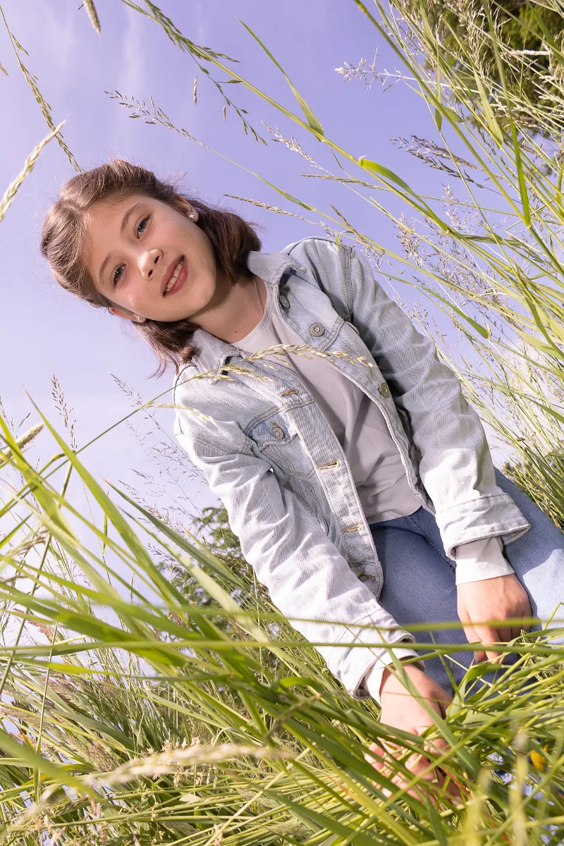 Mädchen in Jeansjacke sitzt im hohen Gras, von unten gegen einen fliederfarbenen Himmel fotografiert