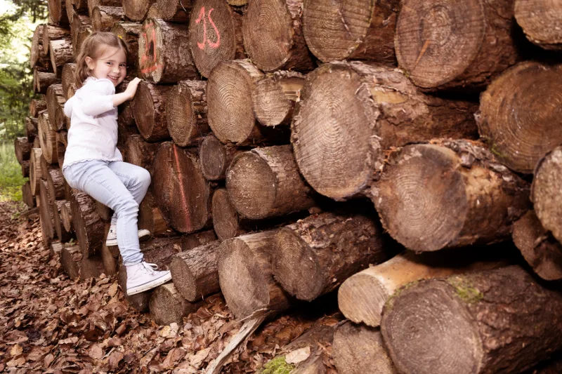 Kleines Mädchen klettert an einem Stapel Baumstämme im Wald und lacht in die Kamera