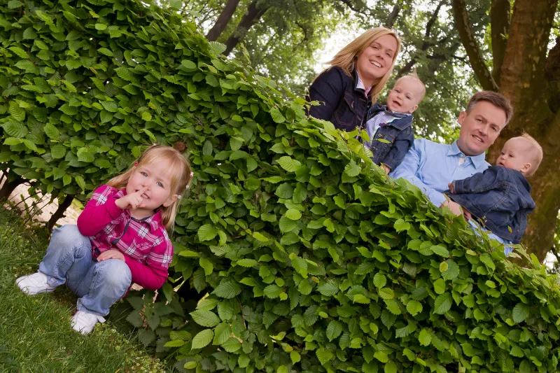 Kleines Mädchen mit Finger vor dem Mund an einer hohen Hecke, dahinter die Eltern mit zwei Kleinkindern
