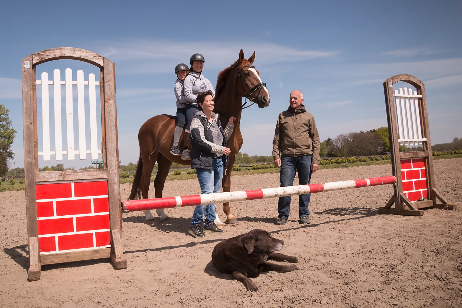Familie auf dem Reitplatz mit Fuchspferd und Hund vor einem rot-weißen Sprunghindernis