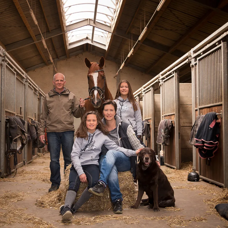 Familie mit Fuchspferd und braunem Labrador im Stallgang, die Töchter in Reiter-Hoodies
