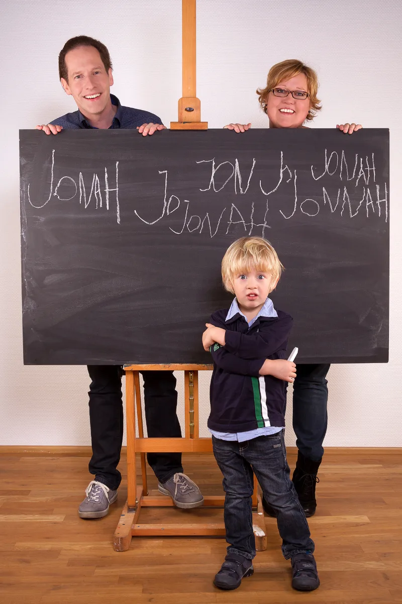 Eltern halten eine Tafel, auf der ein Kindername vielfach in Kreide steht; der Junge davor mit Kreide in der Hand