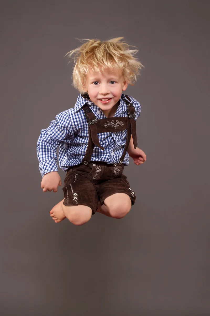 Blonder Junge in Tracht springt barfuß in die Luft, grauer Studiohintergrund