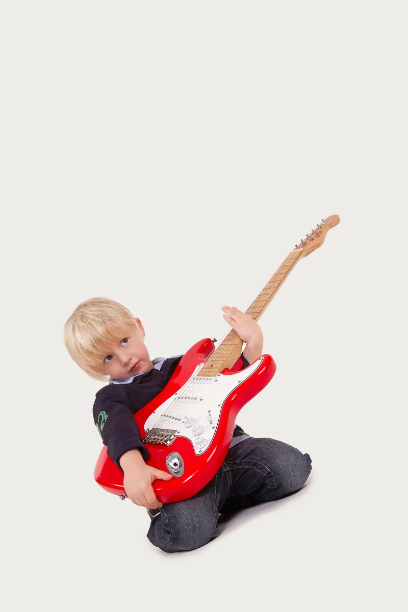 Blonder Junge kniet und umarmt eine rote E-Gitarre vor hellem Hintergrund