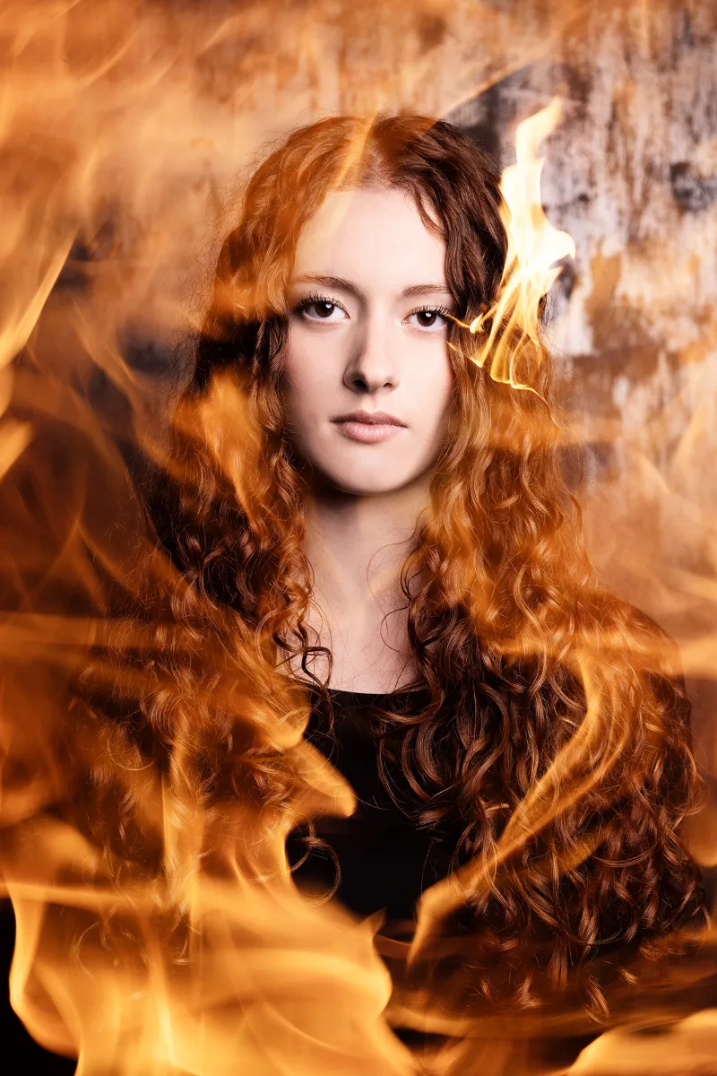 Kreatives Porträt einer jungen Frau mit langem lockigem Haar, umspielt von echten Flammen