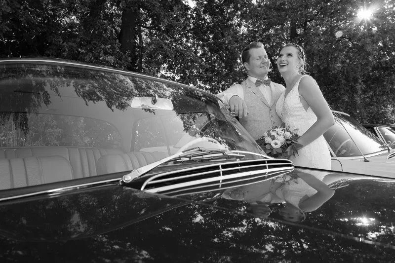 Brautpaar in Schwarz-Weiß am offenen Oldtimer-Cabrio, lachend, Sonne im Gegenlicht