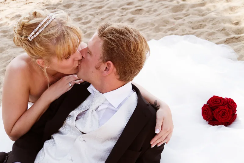 Brautpaar am Strand im Sand, Kuss in Großaufnahme, Brautstrauß mit roten Rosen daneben
