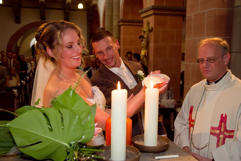 Brautpaar zündet zusammen mit dem Priester eine Hochzeitskerze an, im Vordergrund Monstera-Blätter