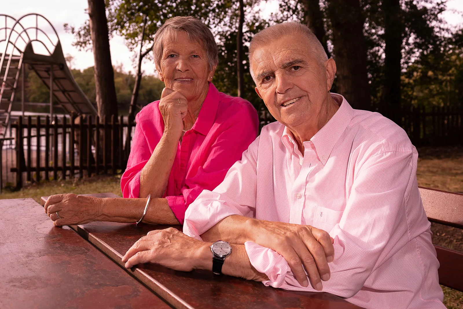Älteres Paar in Rosa und Pink an einem Holztisch im Park, dahinter ein See