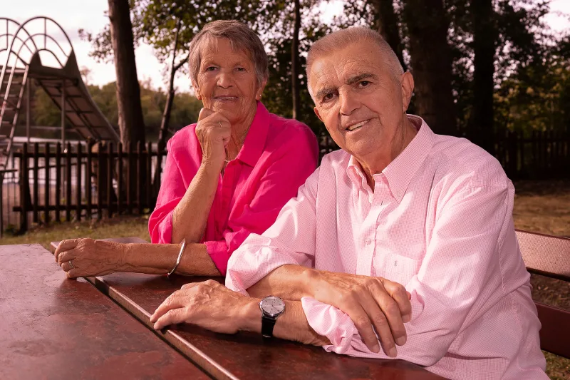 Älteres Paar in Rosa und Pink an einem Holztisch im Park, dahinter ein See