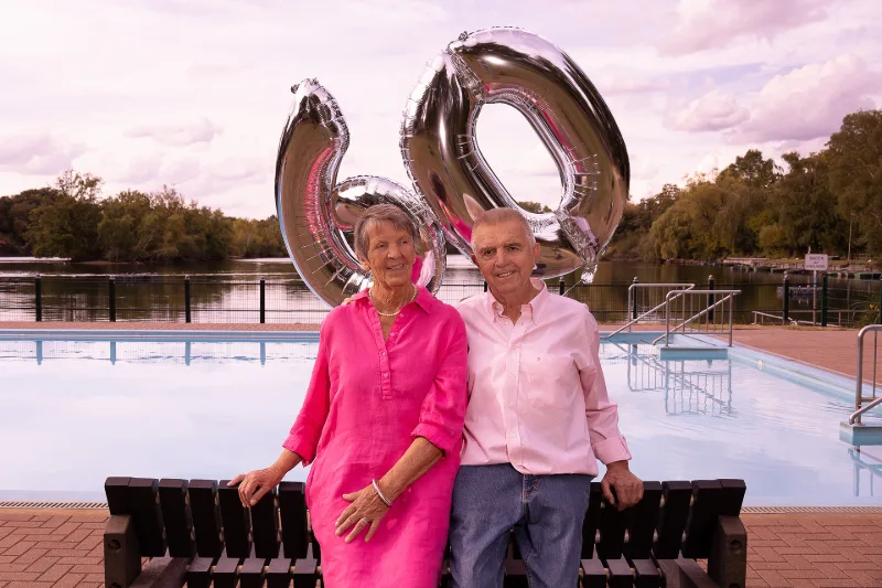 Ehepaar in Pink und Rosa auf einer Holzbank am Freibad-Becken, dahinter silberne Helium-Ballons in Form der Zahl 60