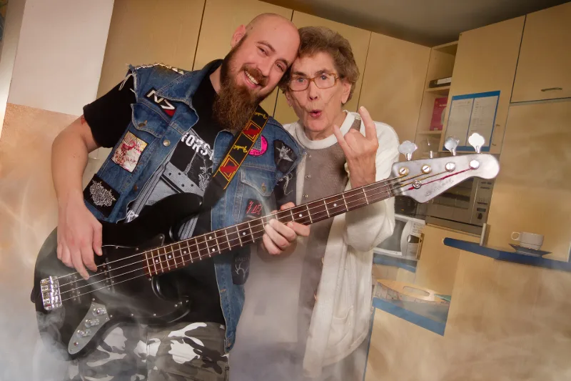 Eine ältere Bewohnerin und ein junger Heavy-Metal-Fan mit Bassgitarre rocken mit Metal-Geste in einer kleinen Küche, Bühnennebel inklusive